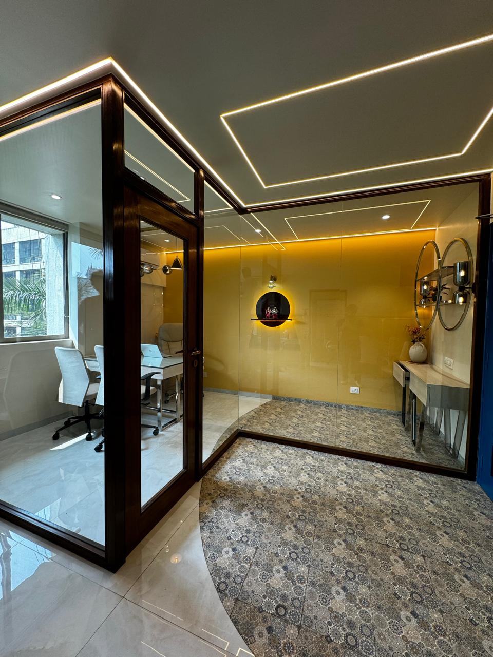 This office showcases a sleek and modern design, featuring glass partitions framed in rich wood, highlighted by recessed and LED strip lighting. The elegant transition from marble to patterned tile flooring, the warm yellow accent wall, and a striking navy blue door create a sophisticated and inviting workspace. The contemporary decor, including a circular shelf and stylish furniture, adds a touch of personality and style to the office